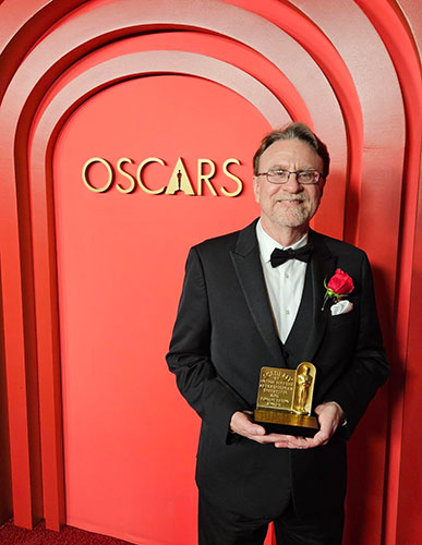 Man dressed in a tuxedo holding a gold award standing in front of a red wall with the word &ldquo;Oscars&rdquo; on it. 