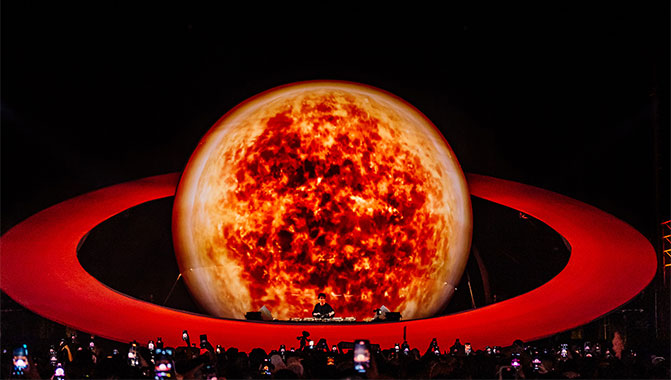 A crowd in front of a large projection mapped replica of Saturn, with a DJ between the planet and its ring.