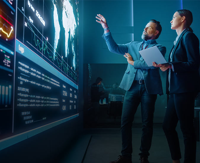 Two people gesturing to a map and data on a large video wall.