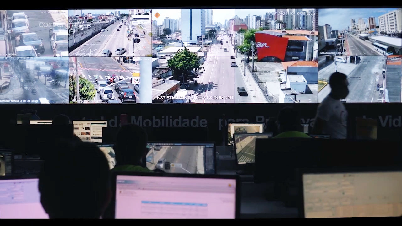 Large LCD video wall displaying roads and cars. In front, people are working at desks with computer screens.