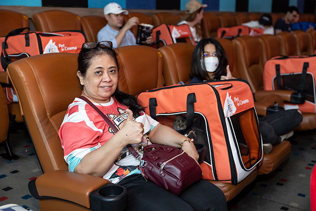 Two women give the thumbs up sign while sitting in beige seats in a cinema with a cat inside a carrier between them. 
