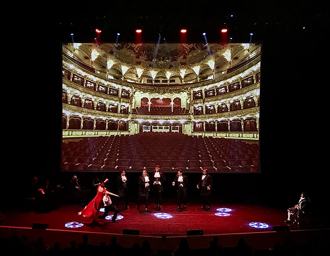 Performers on a dark stage with a projected image of an interior of a theatre in the background.