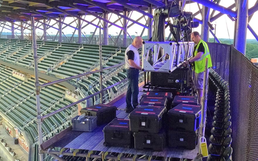 Two people stand on scaffolding to rig projectors in a stadium.