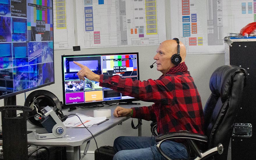 A man with headphones sits at a desk and points to a large display monitor.
