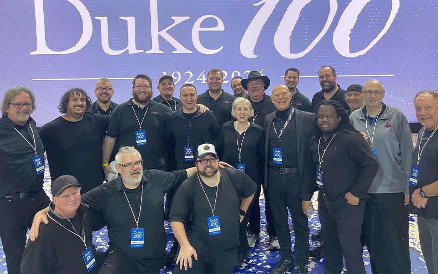 A group of people stand in front of a large sign with the text, &ldquo;Duke 100.&rdquo;