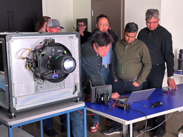 A group of people look at a laptop computer screen on a table beside a large projector. 